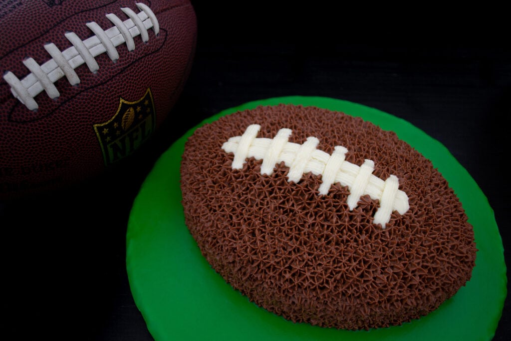 Kuchen in Form eines American Footballs bedeckt mit einer weißen und braunen Schokoladencreme auf einer grünen Kuchenplatte.