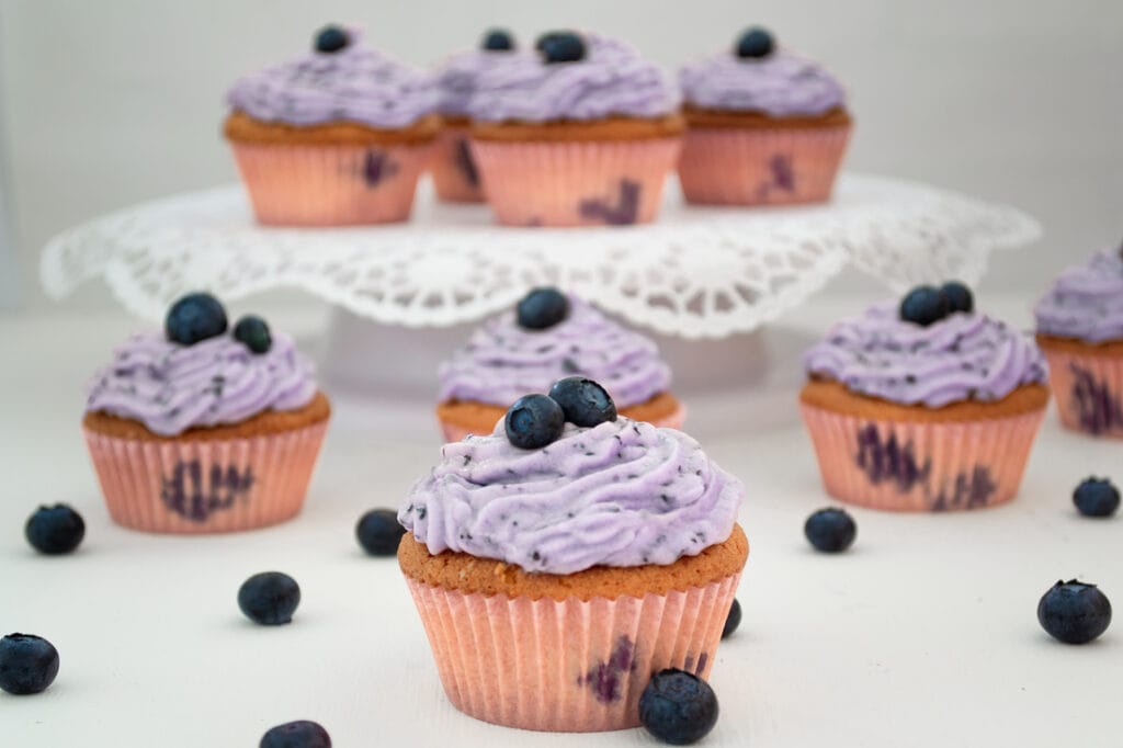 Helle Blaubeer-Cupcakes, bespritzt mit einem leicht lilafarbenem Frosting mit kleinen Blaubeerstückchen. Auf dem Frosting und um die Cupcakes herum liegen frische Blaubeeren.