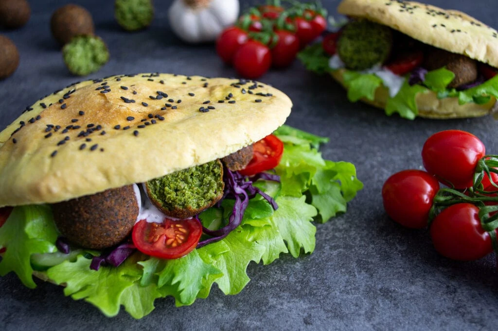 Zwei Falafel-Döner liegen auf einem dunklen Untergrund. Die Pita-Taschen der Falafel-Döner sind mit schwarzem Sesam bestreut und gefüllt mit frischem, grünem Salat, knalligen, roten Tomatenscheiben, lilafarbenem Rotkohlstreifen, runden Falafeln und einer weißen Knoblauchsauce.