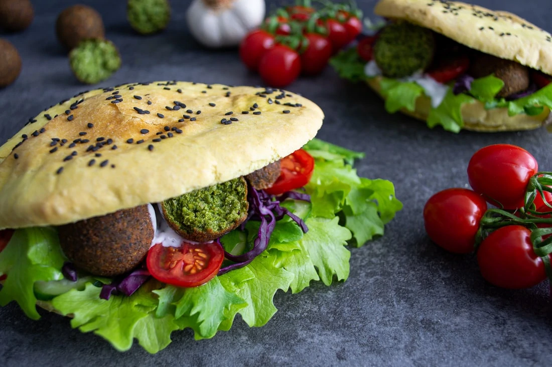 Zwei Falafel-Döner liegen auf einem dunklen Untergrund. Die Pita-Taschen der Falafel-Döner sind mit schwarzem Sesam bestreut und gefüllt mit frischem, grünem Salat, knalligen, roten Tomatenscheiben, lilafarbenem Rotkohlstreifen, runden Falafeln und einer weißen Knoblauchsauce.