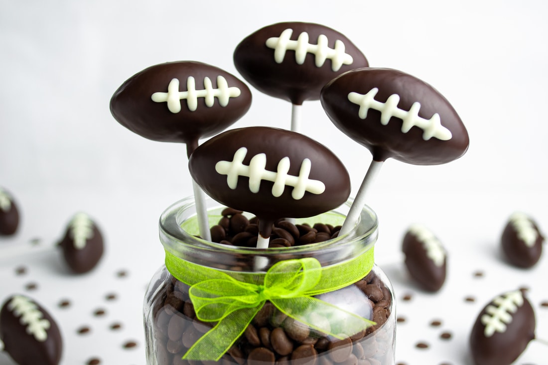 Vier Football Cake Pops, überzogen mit dunkler Schokolade und verziert mit einer weißen Schokoladen-Naht, stecken in einem Glas gefüllt mit Schokoladendrops