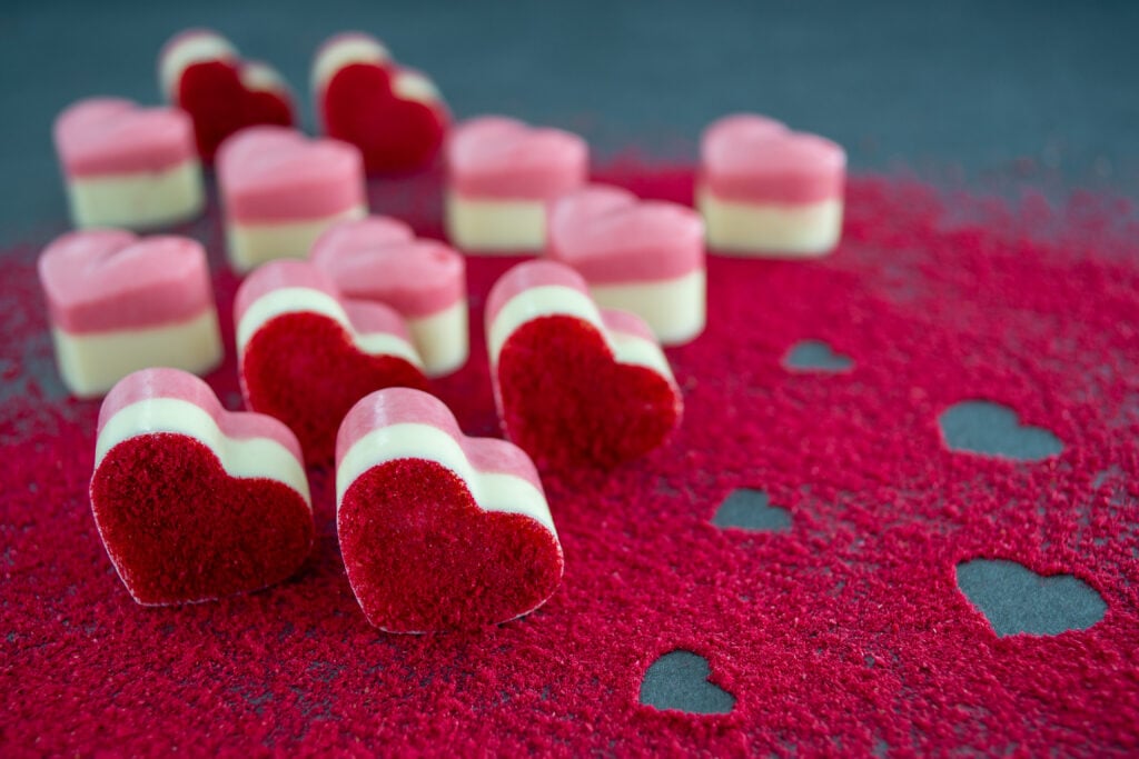 Mehrere Himbeerpralinen aus rosa und weißer Schokolade in Form von kleinen Herzen liegen auf einer mit rotem Himbeerpulver bestreuten, dunklen Platte. Im Himbeerpulver sind kleine freie Stellen in Herzform.