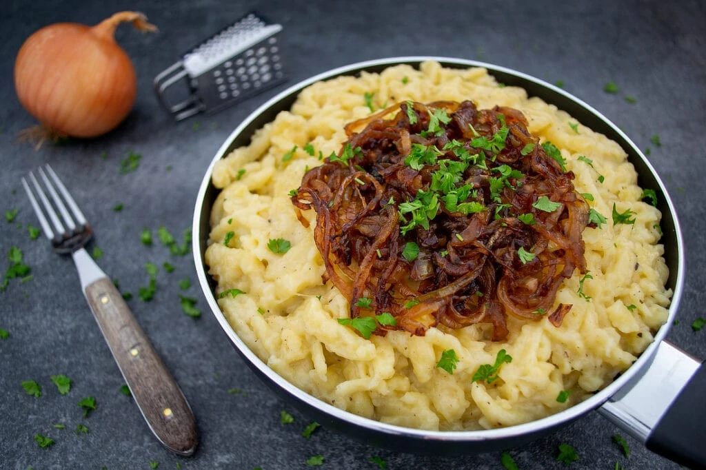 Käsespätzle mit karamellisierten Zwiebeln angerichtet in einer Pfanne garniert mit gehackter Petersilie.