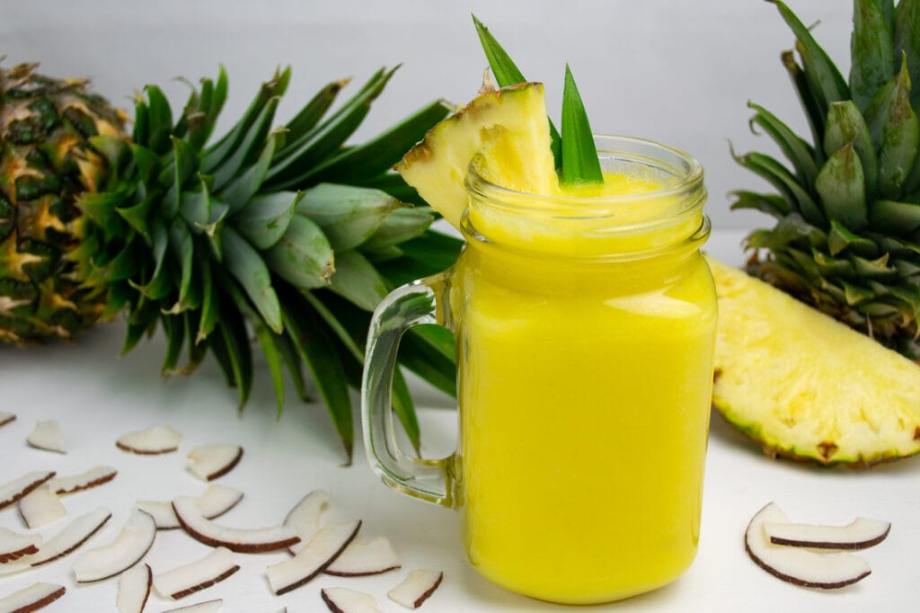 Im Vordergrund des Bildes steht ein gelber Piña Colada Smoothie in einem Glas mit Henkel. Im Hintergrund steht eine ganze und eine angeschnittene Ananas und um das Glas herum liegen Kokos-Chips.