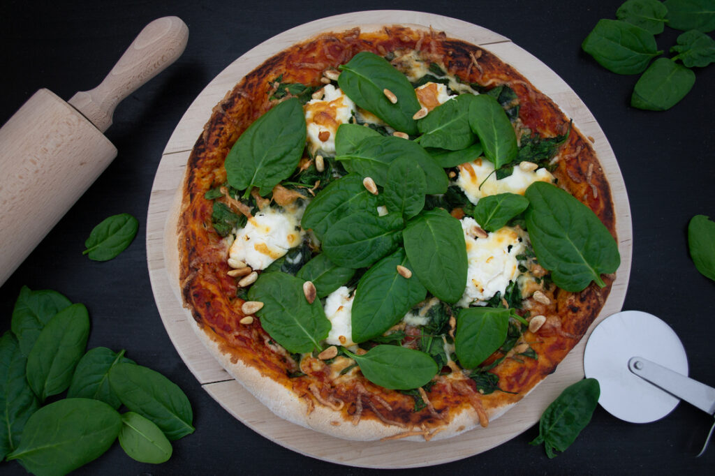 Eine knusprig gebackene Pizza belegt mit Ricotta und frischem Spinat, überbacken mit Käse, liegt auf einem hellen Holzbrett.