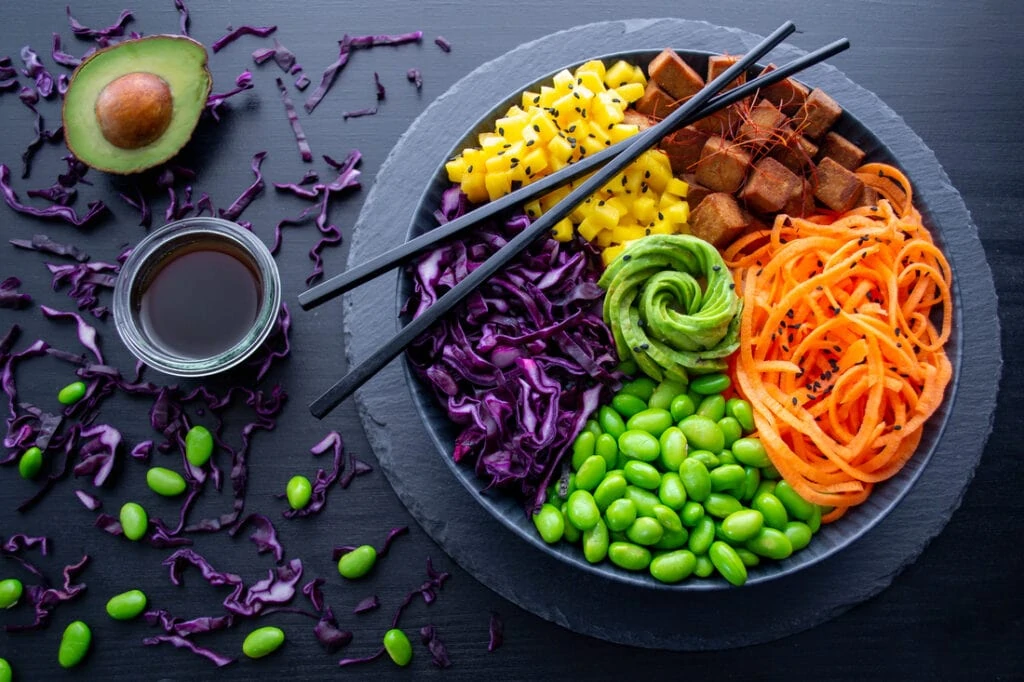 In einem schwarzen, tiefen Teller ist eine Teriyaki Poke Bowl angerichtet. Sie besteht aus orangenen Karottenstreifen, lilafarbenem Rotkraut, braun gebratenen Tofuwürfeln, gelben Mangowürfeln und grünen Edame. Der Blickfang in der Mitte der Teriyaki Poke Bowl ist eine Rose aus Avocado.