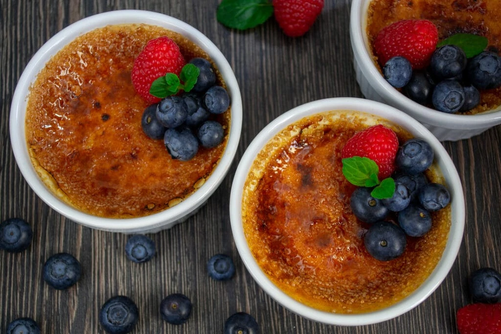 Drei Crème Brûlée in weißen Souffléförmchen, garniert mit Blaubeeren, Minze und Himbeeren
