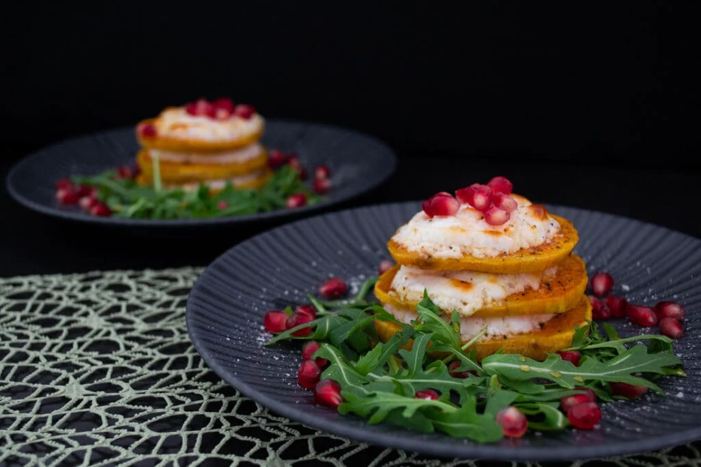 Süßkartoffel-Ziegenkäse-Türmchen auf schwarzem Teller mit Rucola und Granatapfelkernen