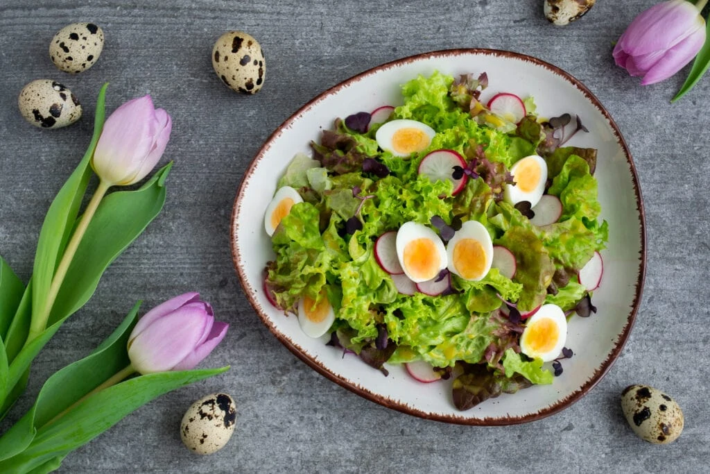 Ein bunter Frühlingssalat mit Wachteleiern und Radieschen, angemacht mit einem cremigen, gelben Honig-Senf Dressing ist auf einem weißen Teller mit braunen Rand angerichtet. Um den Teller herum liegen drei rosafarbene Tulpen und sechs ganze Wachteleier.