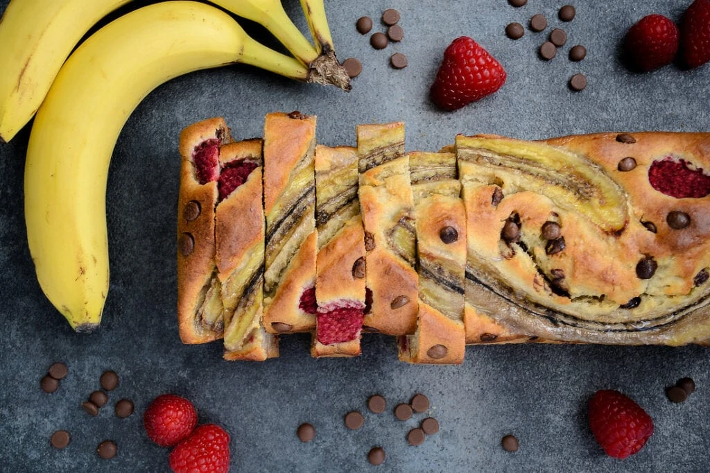 Das Himbeer-Schokoladen Bananenbrot liegt aufgeschnitten auf einem dunklen Untergrund. Um das Bananenbrot herum liegen frische Himbeeren, Schokotröpfchen und Bananen.