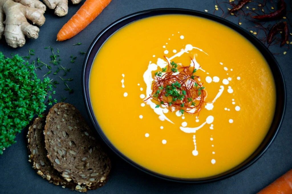 Eine orangene Karottensuppe mit Ingwer und Chili serviert in einem tiefen, ovalen Teller. Um den Teller mit der Karottensuppe mit Ingwer und Chili liegen zwei Scheiben Brot, Chilis, Karotten, Kresse und Ingwerwurzeln.