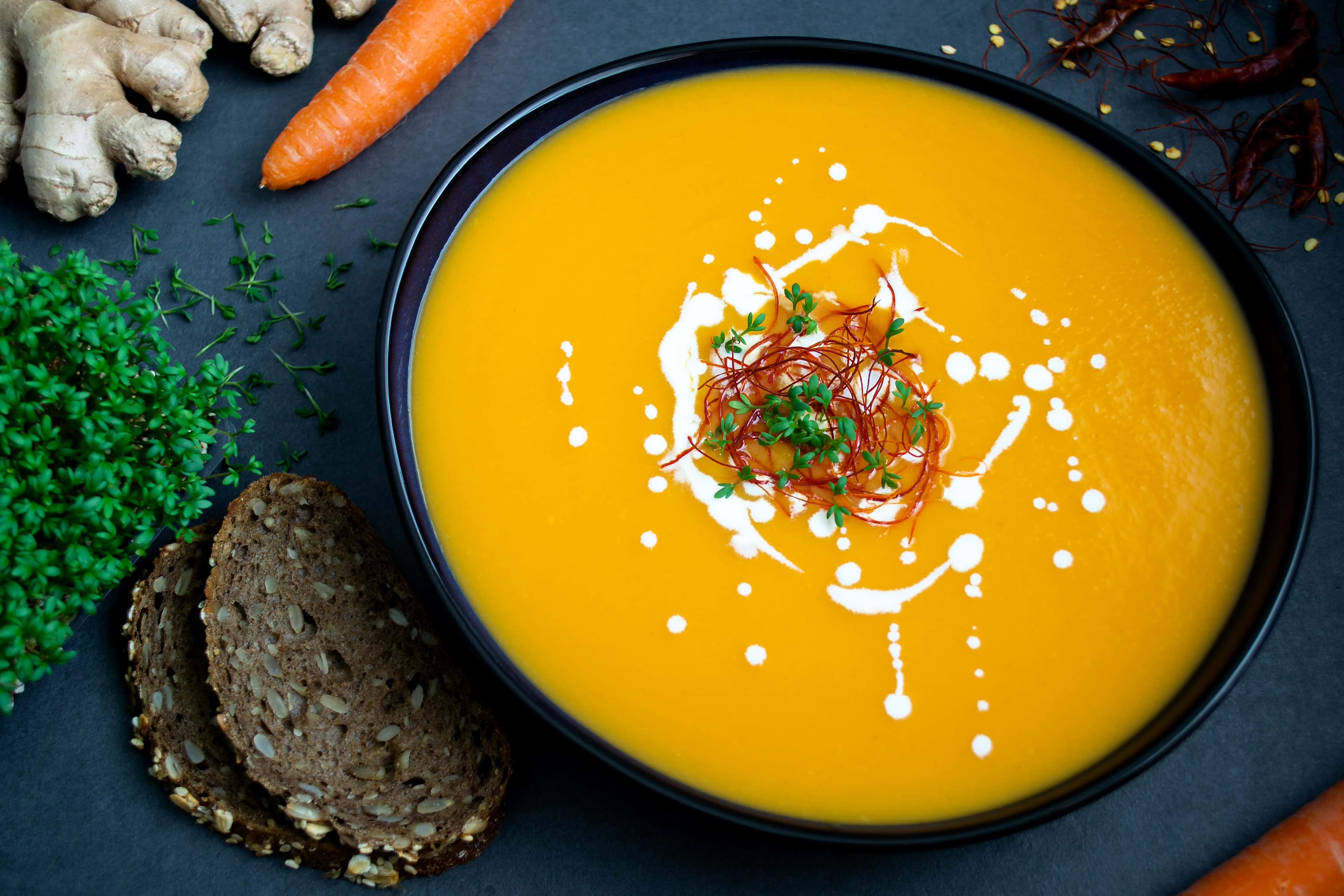 Eine orangene Karottensuppe mit Ingwer und Chili serviert in einem tiefen, ovalen Teller. Um den Teller mit der Karottensuppe mit Ingwer und Chili liegen zwei Scheiben Brot, Chilis, Karotten, Kresse und Ingwerwurzeln.
