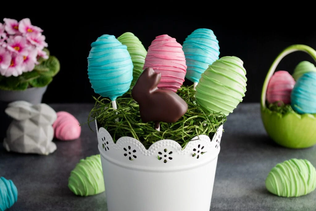 Im Vordergrund des Bildes stecken bunte Oster Cake Pops in einem weißen Topf verziert mit Ostergras. Im Hintergrund liegen weitere Cake Pops, eine rosa Blume und ein kleiner, grauer Osterhase.