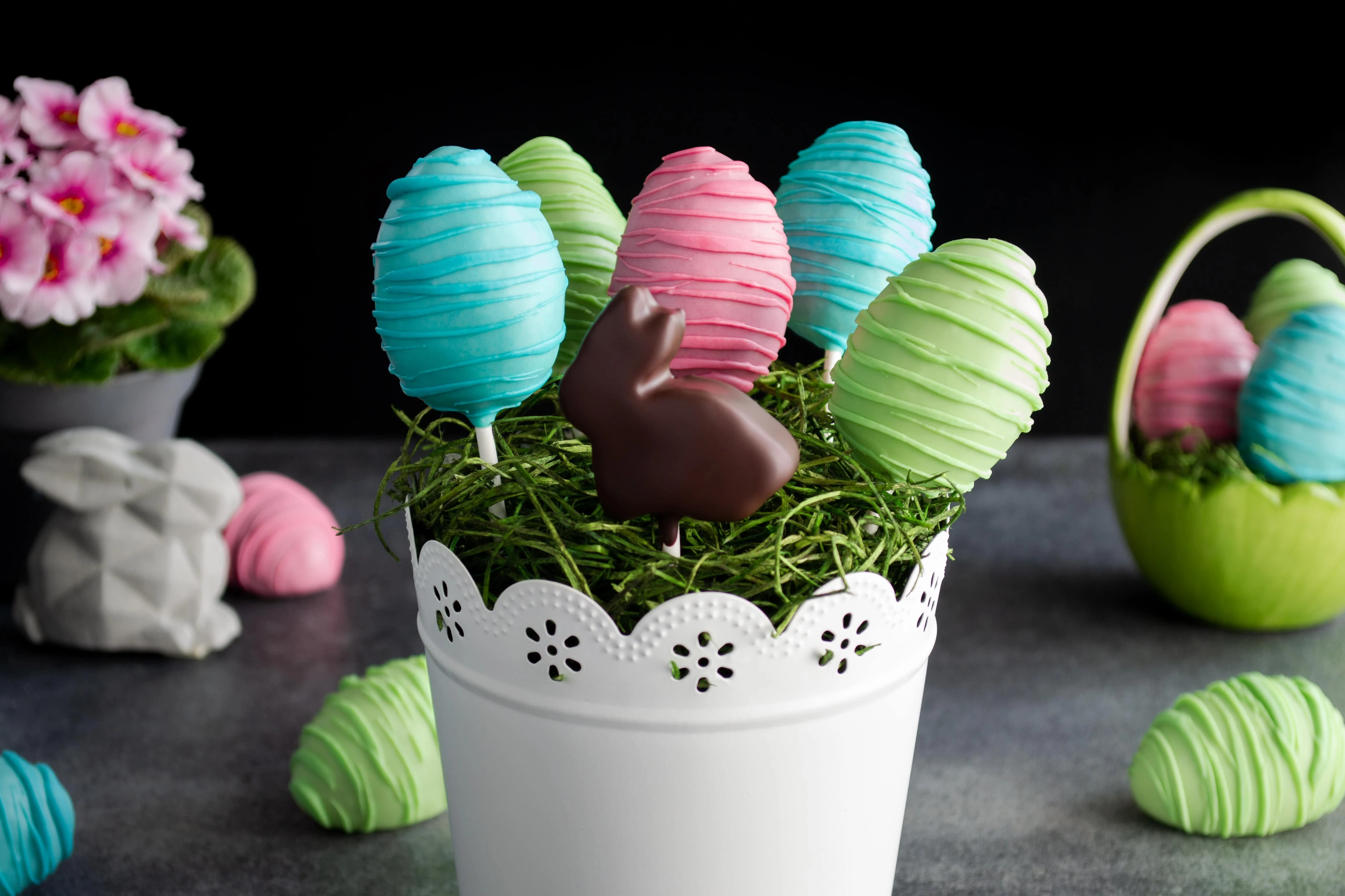 Im Vordergrund des Bildes stecken bunte Oster Cake Pops in einem weißen Topf verziert mit Ostergras. Im Hintergrund liegen weitere Cake Pops, eine rosa Blume und ein kleiner, grauer Osterhase.