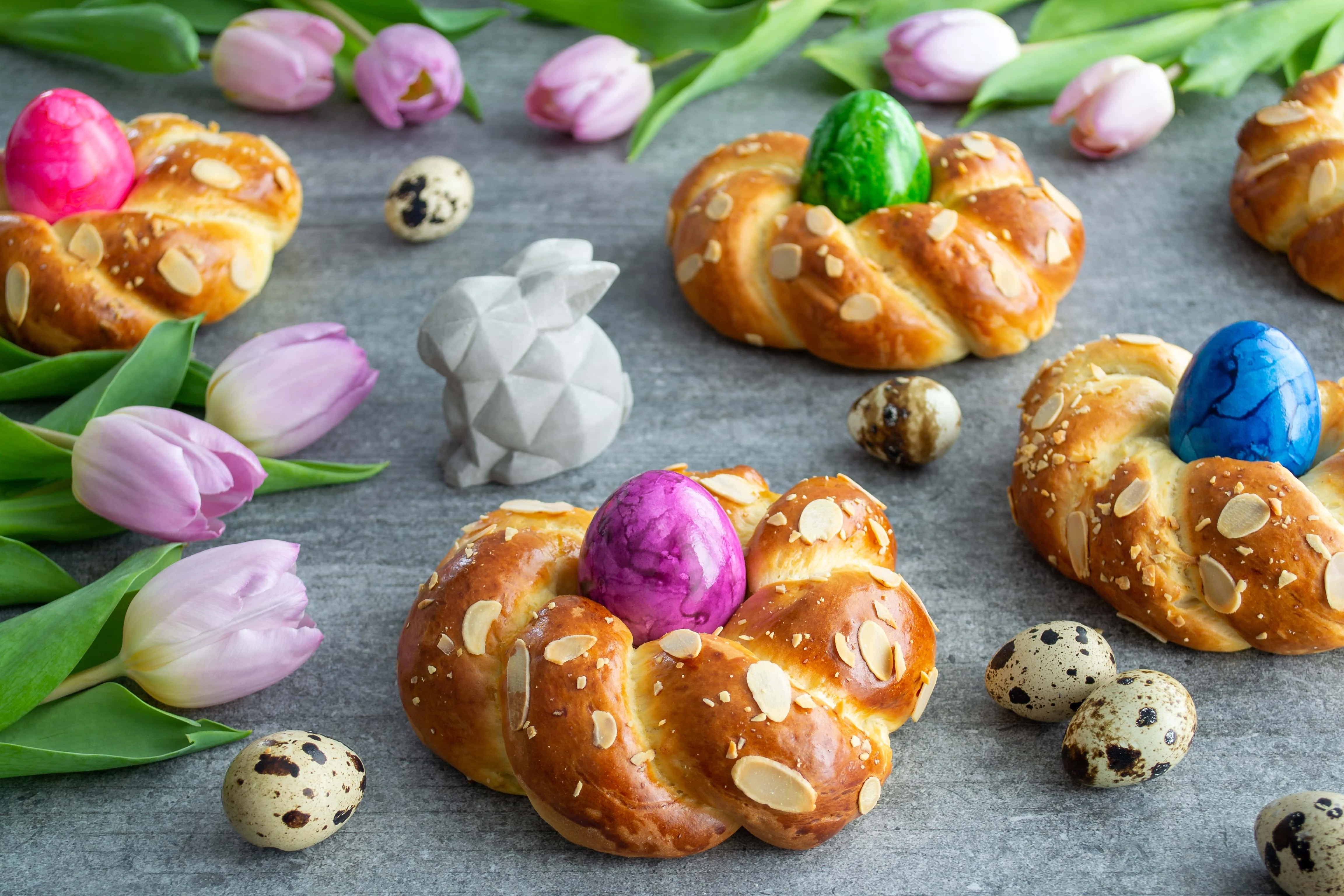 Vier Osterkränze aus Hefeteig, gefüllt mit jeweils einem bunten Osterei, liegen auf einem grauen Untergrund. Neben den Osterkränzen aus Hefeteig liegen rosafarbene Tulpen, kleine Wachteleier und eine Osterhasenfigur.