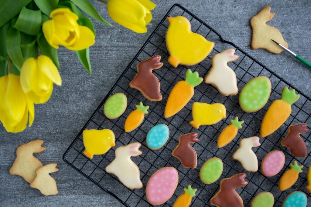 Auf einem Abkühlgitter liegen bunte, verzierte Osterplätzchen mit Zuckerguss. Die Osterplätzchen haben die Form von Osterhasen, Küken, Ostereiern und Karotten. An der Seite steht ein Blumenstrauß mit gelben Tulpen.