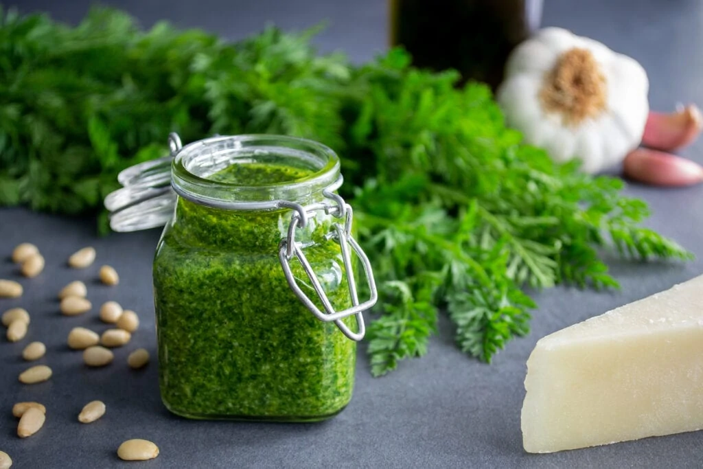Ein grünes Pesto aus Karottengrün abgefüllt in einem Glas steht im Vordergrund. Neben dem Pesto aus Karottengrün liegen Pinienkerne, Knoblauch, Parmesan und Karottengrün.