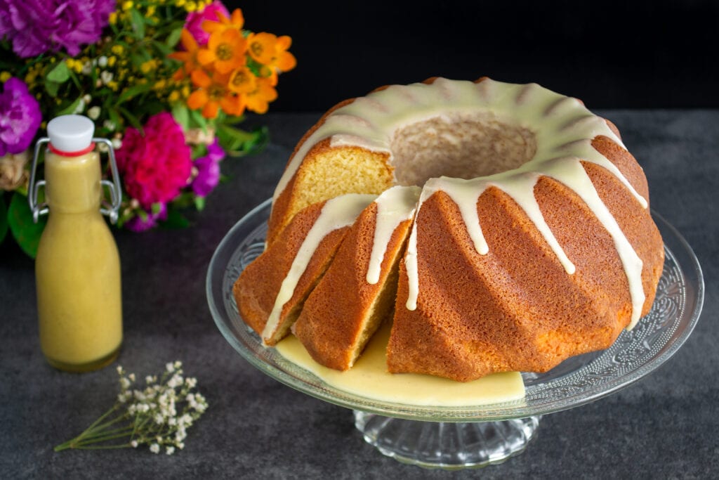 Ein saftiger Eierlikörkuchen überzogen mit einer hellen Glasur liegt auf einer Kuchenplatte. Hinter dem saftigen Eierlikörkuchen steht ein bunter Blumenstrauß und eine Bügelflasche gefüllt mit gelbem Eierlikör.