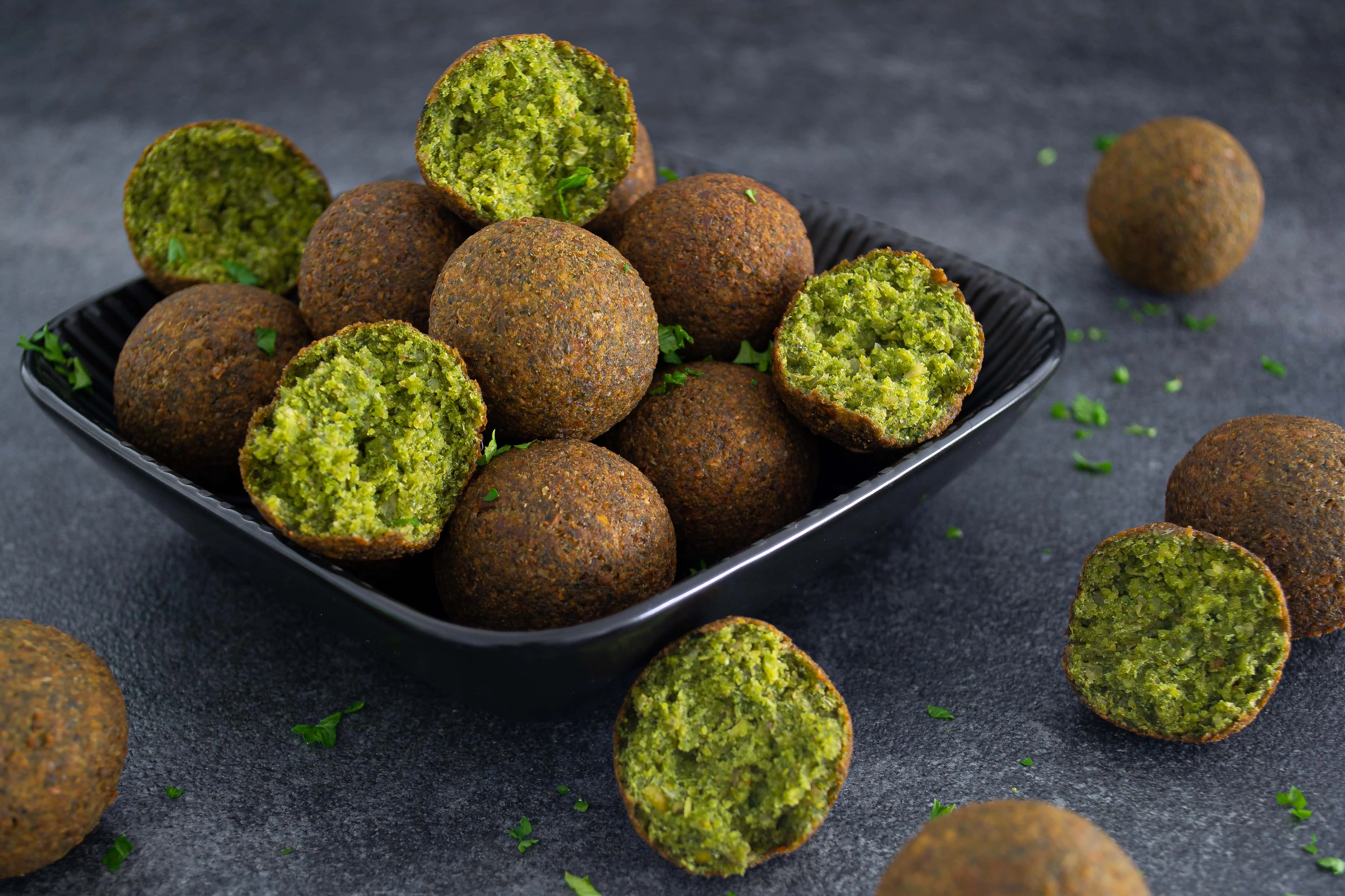 Mehrere Spinat-Falafel, mit einer gold-braunen Kruste, liegen in einer schwarzen, eckigen Schale. Drei der Spinat-Falafel sind halbiert und zeigen den grünen Kern im Inneren.
