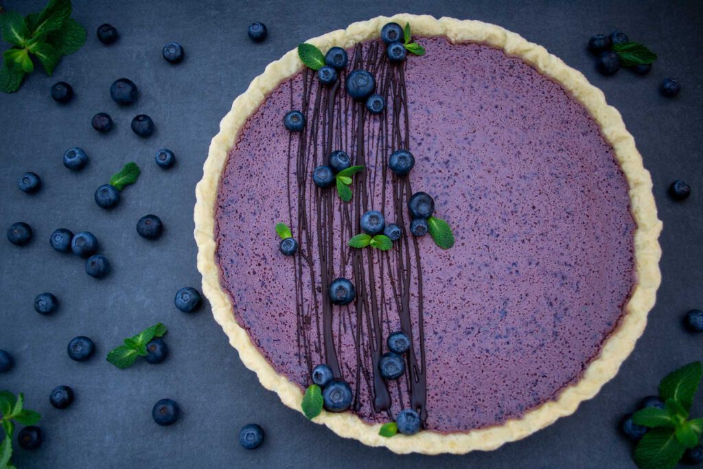 Auf einem dunklen Untergrund liegt eine Blueberry Cheesecake Tarte. Die lila Blaubeer-Füllung der Tarte ist mit mehreren Streifen Schokolade, Blaubeeren und frischer Minze garniert. Neben der Blueberry Cheesecake Tarte liegen weitere Blaubeeren.
