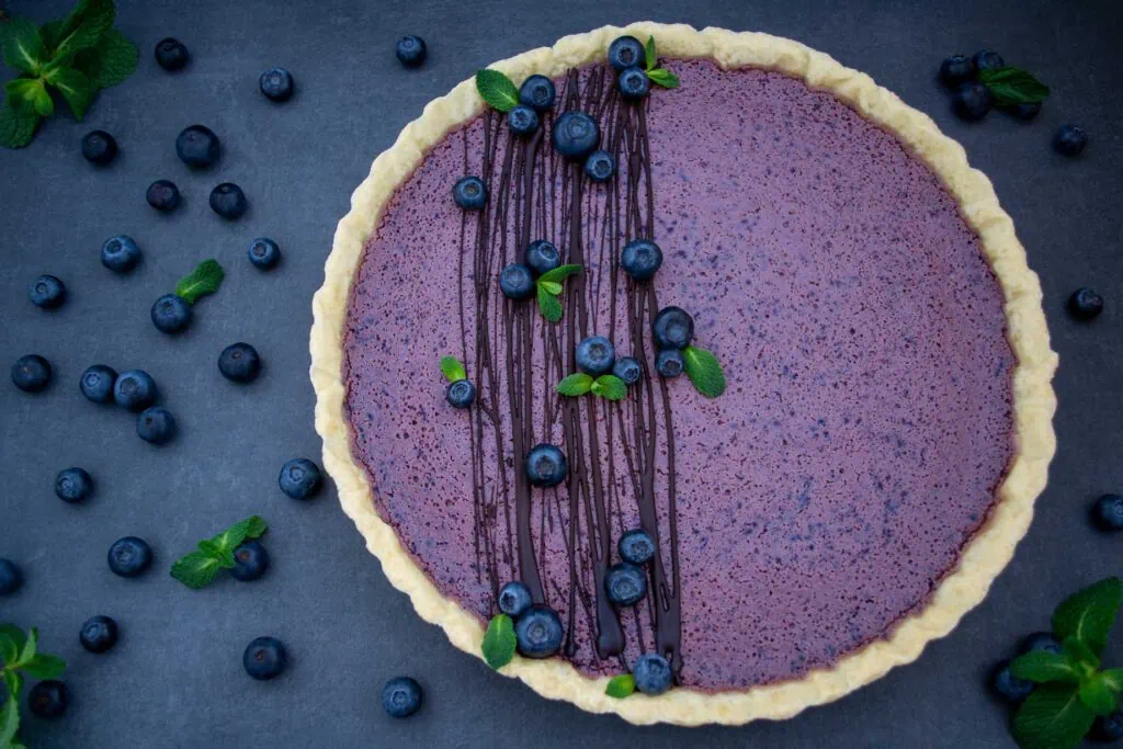 Auf einem dunklen Untergrund liegt eine Blueberry Cheesecake Tarte. Die lila Blaubeer-Füllung der Tarte ist mit mehreren Streifen Schokolade, Blaubeeren und frischer Minze garniert. Neben der Blueberry Cheesecake Tarte liegen weitere Blaubeeren.
