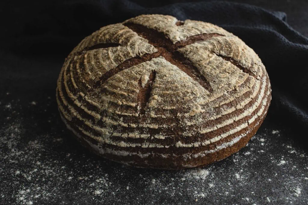 Auf einem dunklen, bemehlten Untergrund liegt ein ganzes Dinkelvollkornbrot mit Sauerteig. Die Oberfläche des Brotes ist kreuzförmig eingeschnitten.