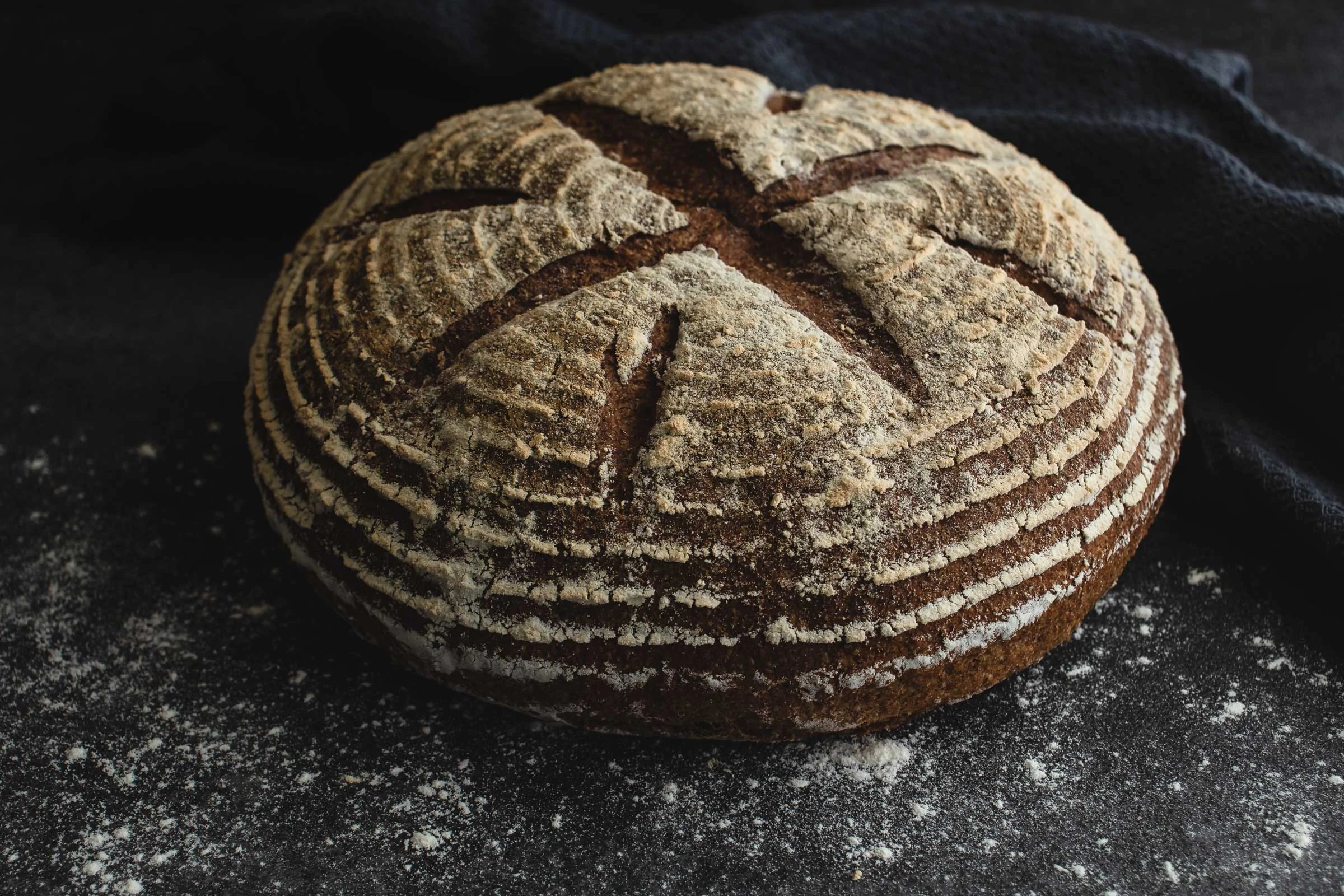 Auf einem dunklen, bemehlten Untergrund liegt ein ganzes Dinkelvollkornbrot mit Sauerteig. Die Oberfläche des Brotes ist kreuzförmig eingeschnitten.