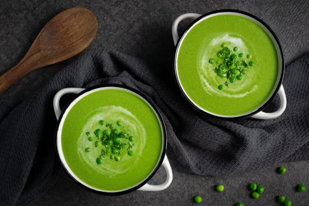 Zwei weiße Porzellantöpfe, gefüllt mit einer grünen Erbsensuppe mit Kokosmilch, stehen auf einem schwarzen Untergrund. Neben der Erbsensuppe liegen ein Geschirrhandtuch, grüne Erbsen und ein Kochlöffel aus Holz.
