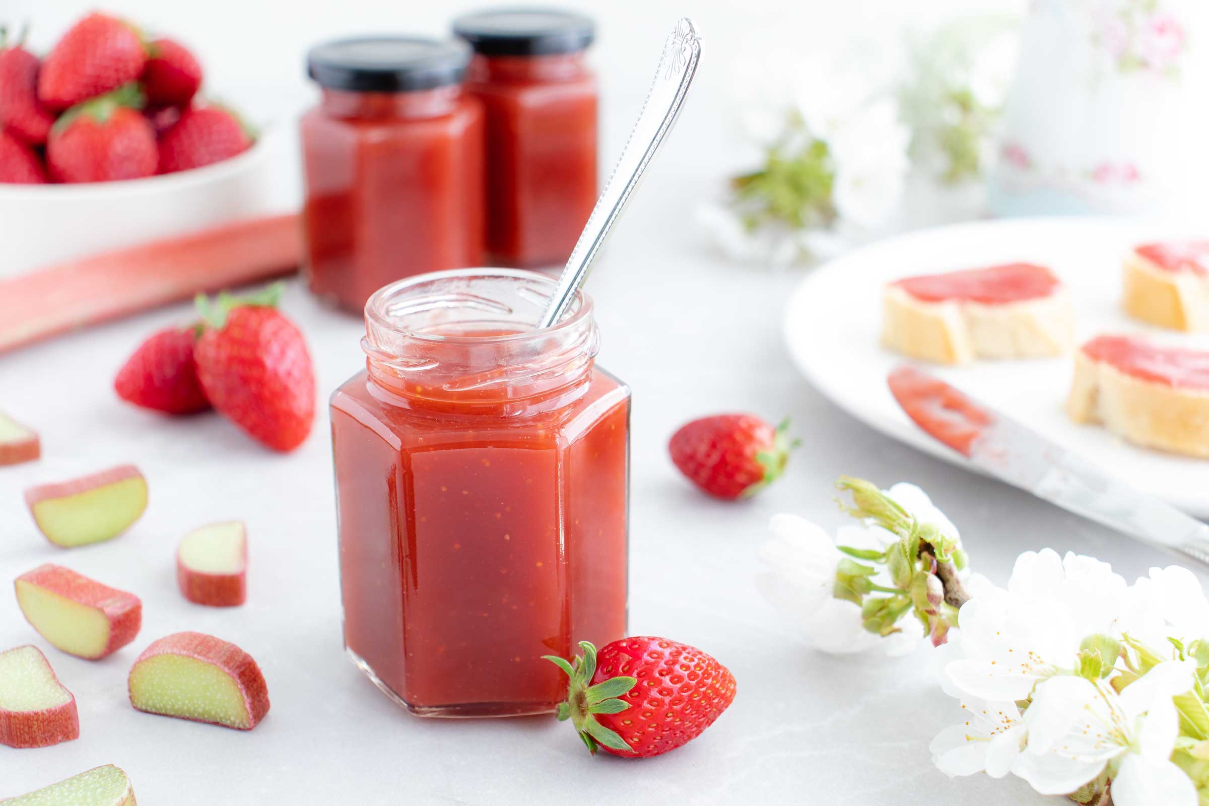 Ein Glas gefüllt mit einer roten Erdbeer-Rhabarber Marmelade steht auf einem weißen Tisch. Neben dem Glas liegen Erdbeeren, Rhabarber und ein Teller auf dem Brote liegen, bestrichen mit der Erdbeer-Rhabarber Marmelade.