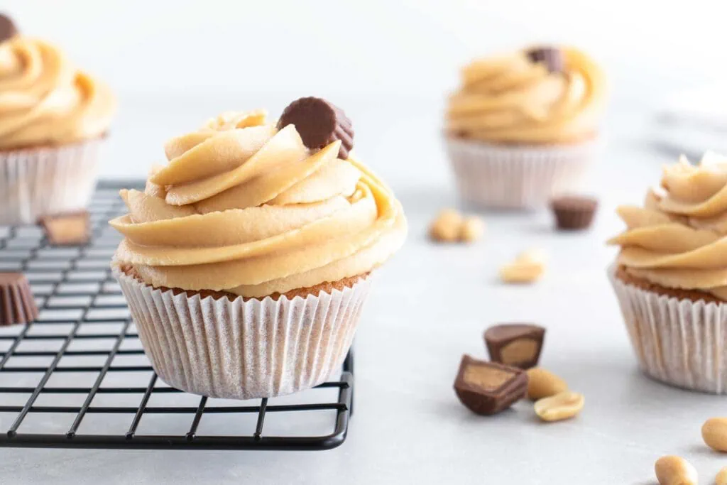 Im Vordergrund steht einer der Erdnussbutter Cupcakes mit einem hellen Frischkäse-Frosting, garniert mit einem Peanut Butter Cup auf einem schwarzen Gitter. Im Hintergrund sind weitere Erdnussbutter Cupcakes und mehrere Peanut Butter Cups zu sehen.