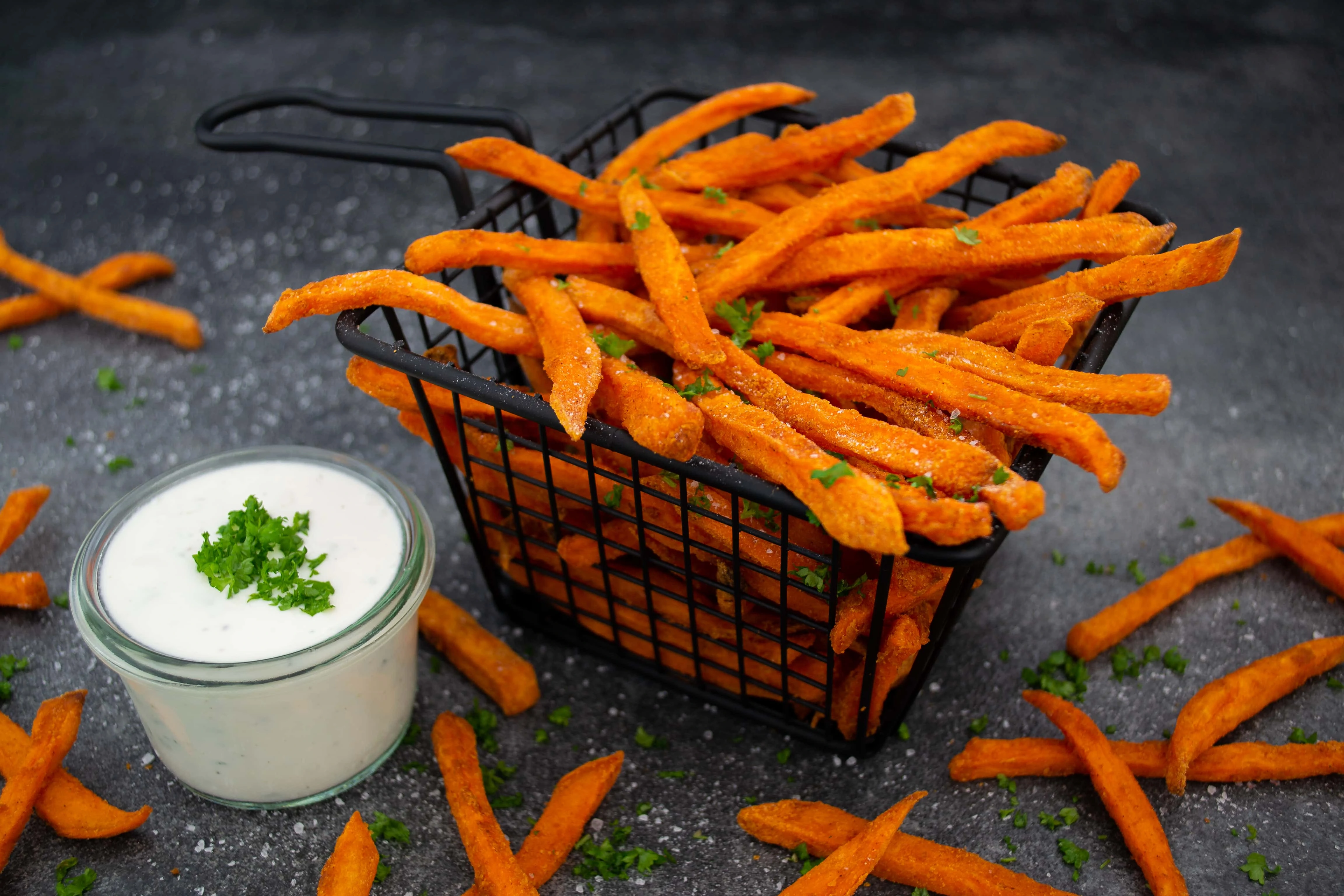 In einem kleinen Frittierkorb liegt eine Portion der knusprigen Süßkartoffelpommes aus dem Backofen. Die Süßkartoffelpommes sind bestreut mit Salz und Petersilie, daneben steht eine Sour Cream als Dip.