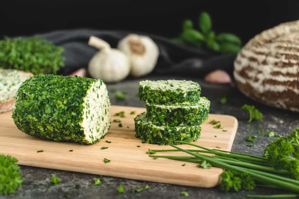 Auf einem kleinen Holzbrett liegt eine in Scheiben geschnittene Kräuterbutter-Rolle. Hinter der Kräuterbutter liegen ein Brotlaib, Knoblauch und verschiedene frische Kräuter.