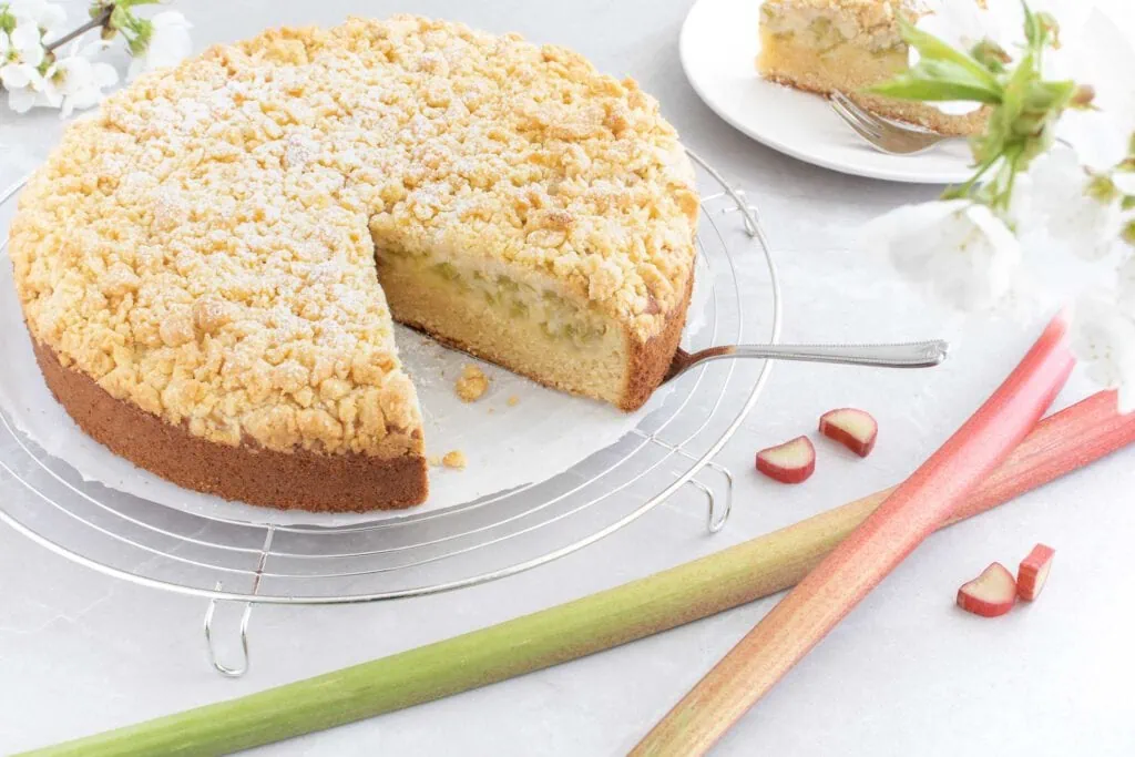 Ein angeschnittener Rhabarberkuchen mit Streuseln, bestäubt mit Puderzucker, liegt auf einem runden Abkühlgitter. Neben dem Rhabarberkuchen liegen, ganze Rhabarberstangen und ein Teller mit einem Kuchenstück.