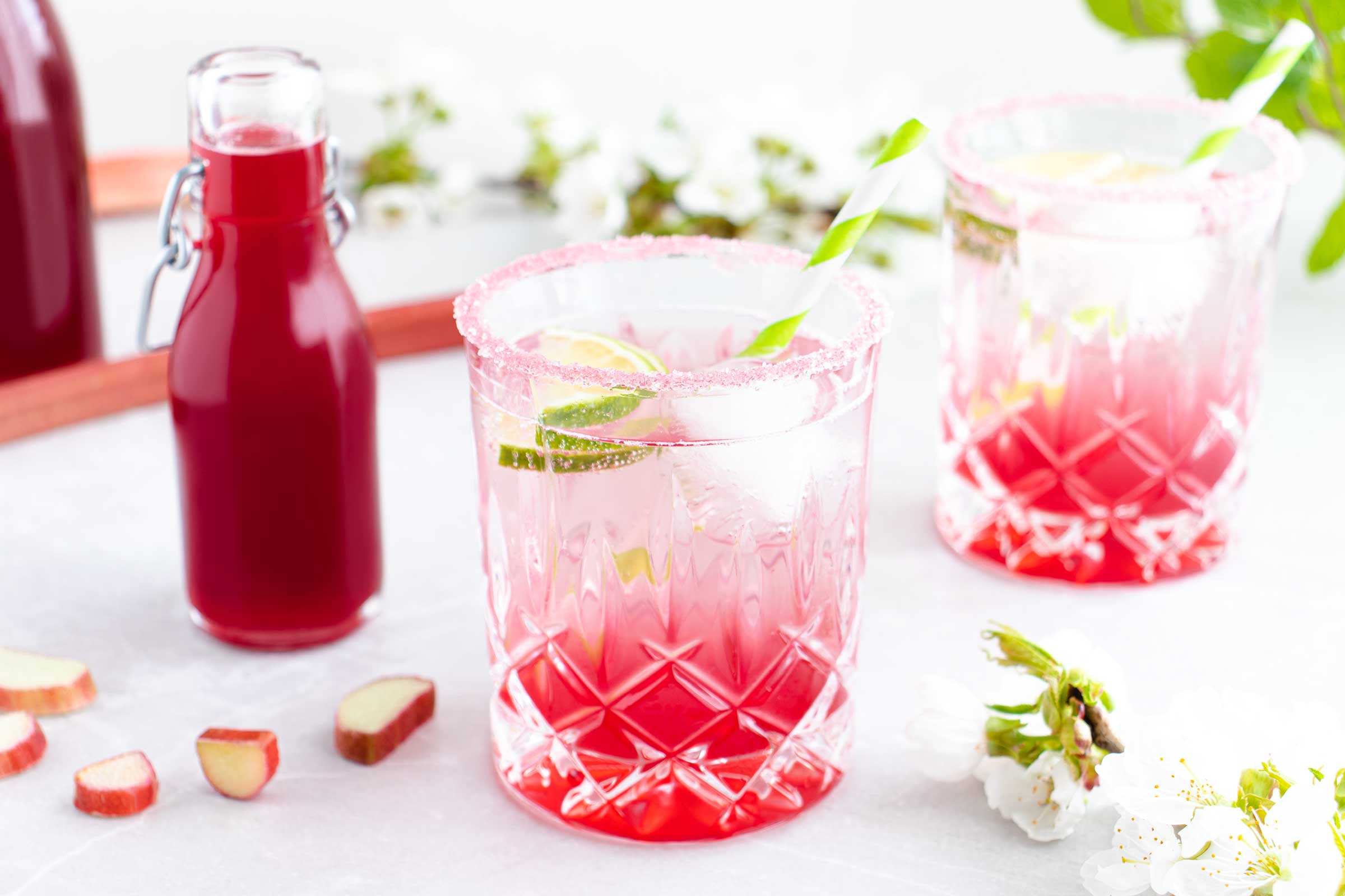 Zwei Gläser gefüllt mit einem erfrischenden Sommergetränk mit selbstgemachten Rhabarbersirup stehen auf einem weißen Tisch. Im Hintergrund stehen zwei Flaschen des roten Rhabarbersirups.