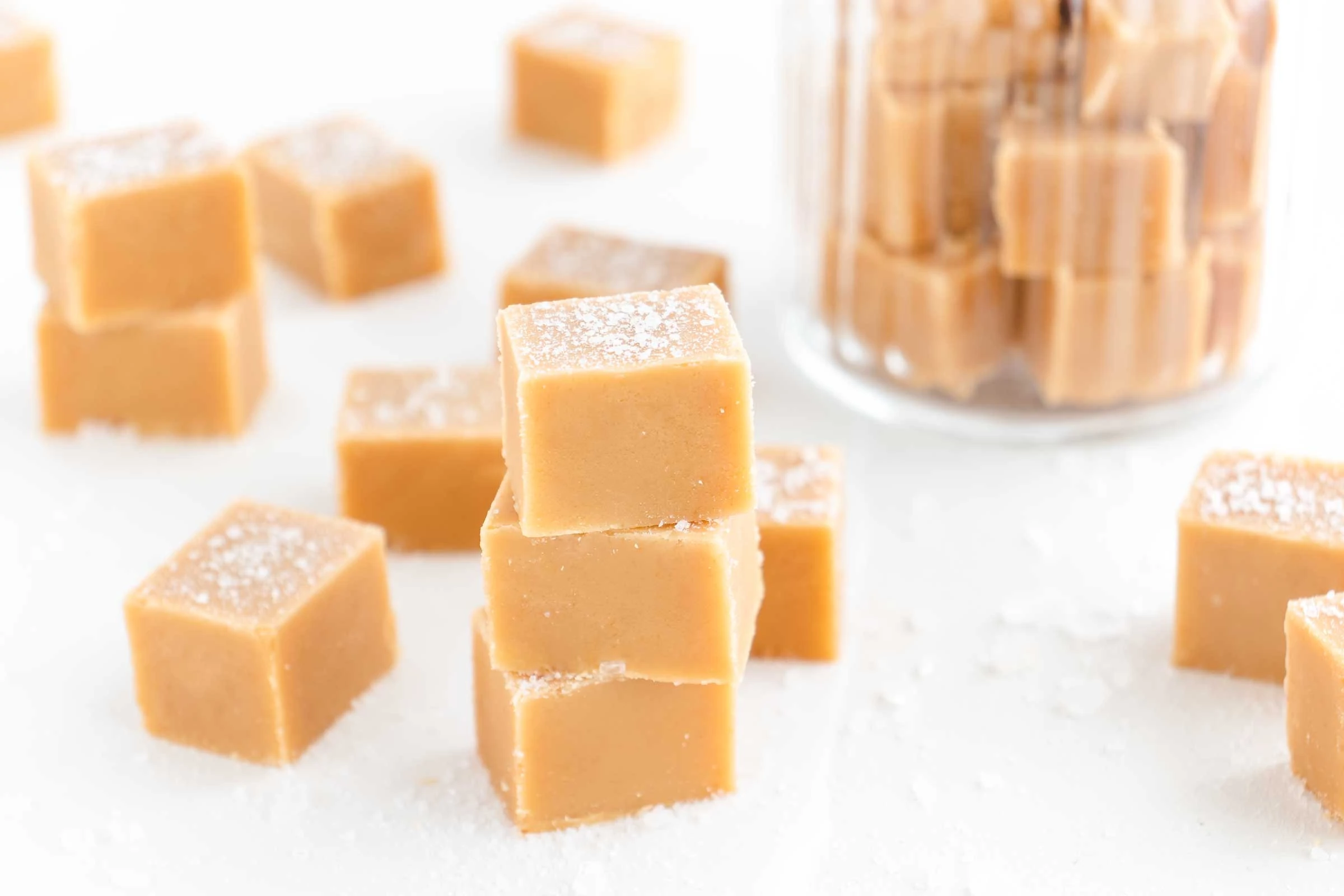 Auf einem weißen, mit grobem Meersalz bestreuten, Untergrund liegen mehrere Würfel des soften Salted Caramel Fudge. Einige der Caramel Fudge Würfel sind aufeinandergestapelt.