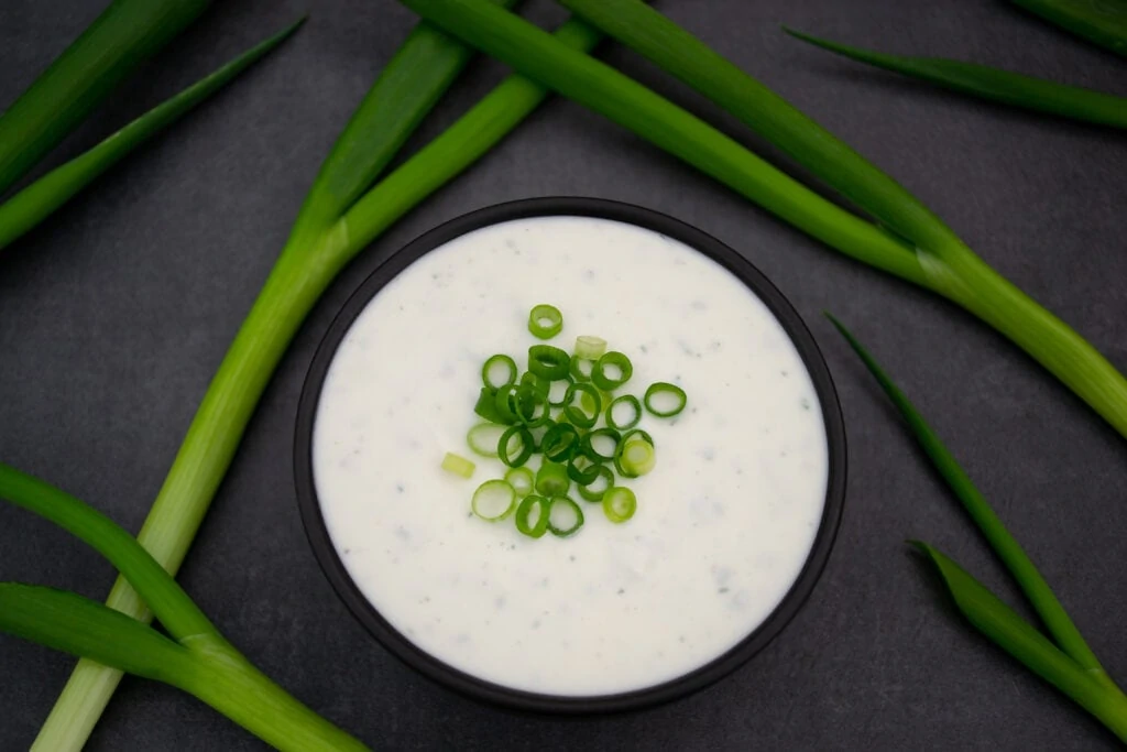 Auf einem dunklen Untergrund steht eine Schale gefüllt mit Sour Cream mit Frühlingszwiebeln. Um die Schale herum liegen mehrere ganze grüne Frühlingszwiebeln.