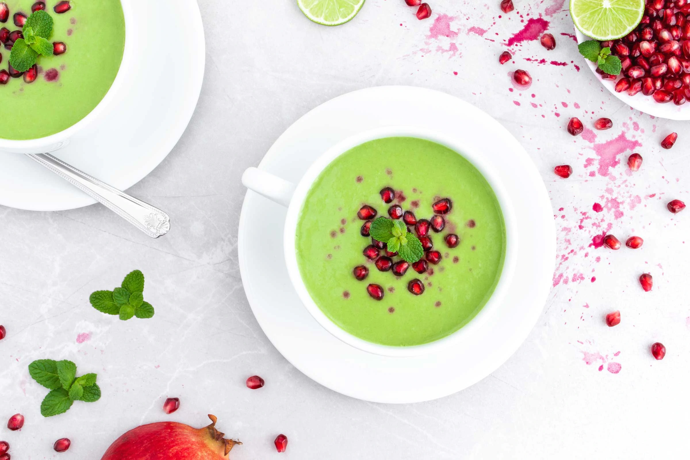 Gurken-Gazpacho garniert mit Granatapfelkernen sind in zwei Suppentassen auf einem hellen Untergrund angerichtet. Neben den Gazpachos liegen Granatapfelkerne und frische Minze.