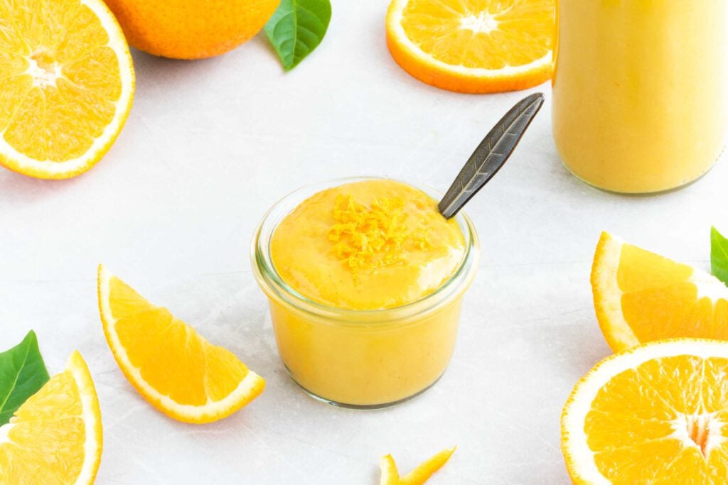 Orangen-Senf-Sauce in einem kleinen Schälchen mit Löffel. Neben der gelben Orangen-Senf-Sauce liegen eine ganze und mehrere geschnittene Orangen.