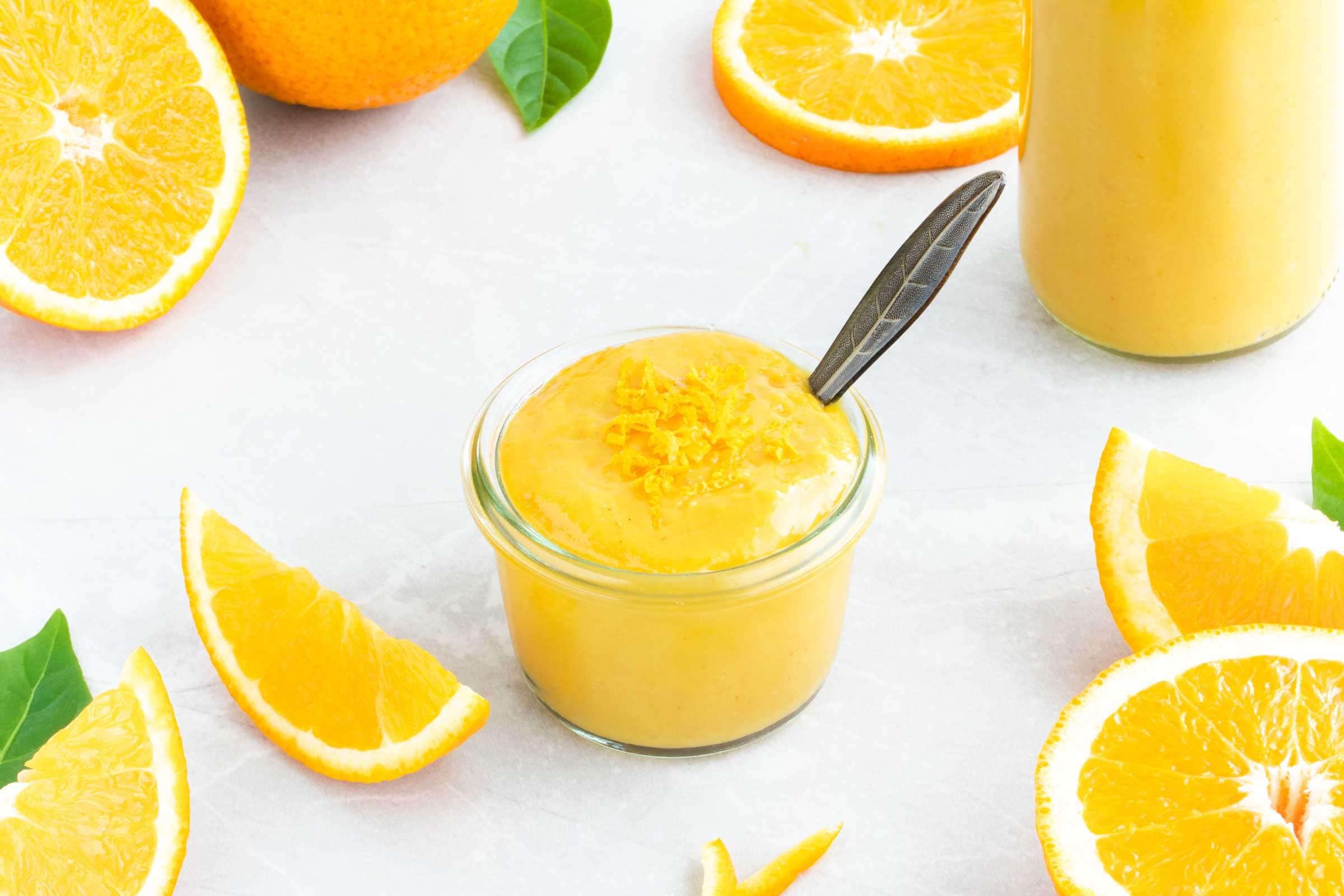 Orangen-Senf-Sauce in einem kleinen Schälchen mit Löffel. Neben der gelben Orangen-Senf-Sauce liegen eine ganze und mehrere geschnittene Orangen.