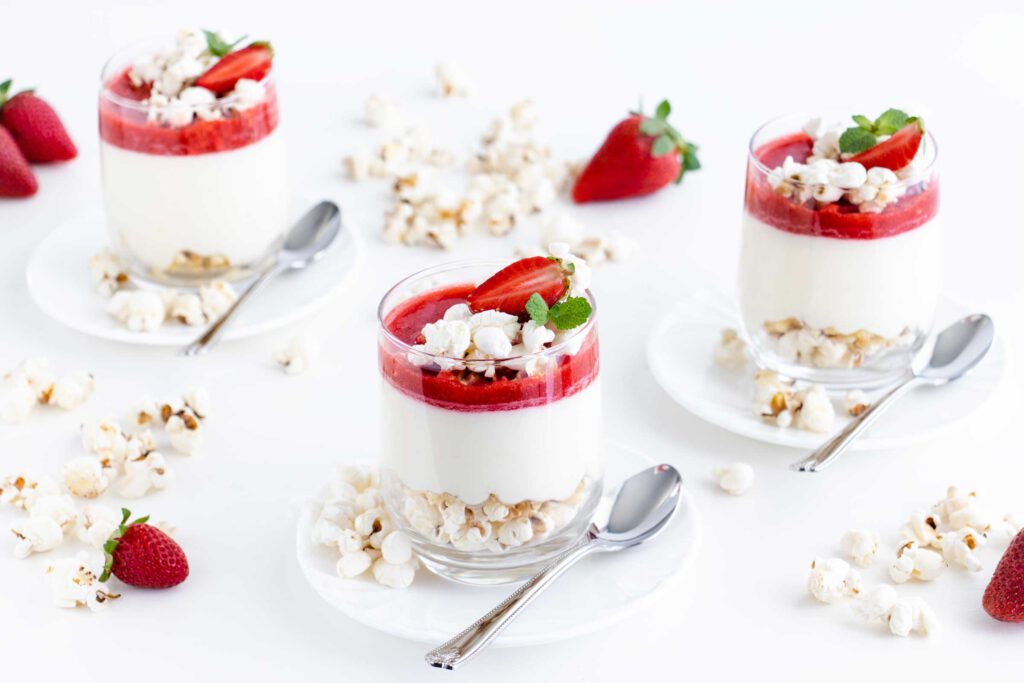 Popcorn Strawberry Cheesecake im Glas auf jeweils einer Untertasse mit Löffel. Neben den Strawberry Cheesecakes liegt Popcorn und frische Erdbeeren.