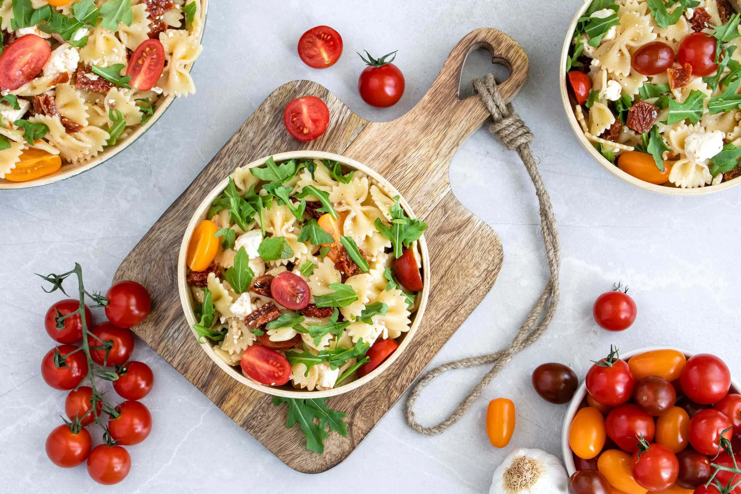 Mediterraner Nudelsalat mit Rucola, Feta und Kirschtomaten in einer Schüssel. Die Schüssel mit dem Nudelsalat steht auf einem Holzbrett mit Griff, daneben liegen mehrere Tomaten und Knoblauch.