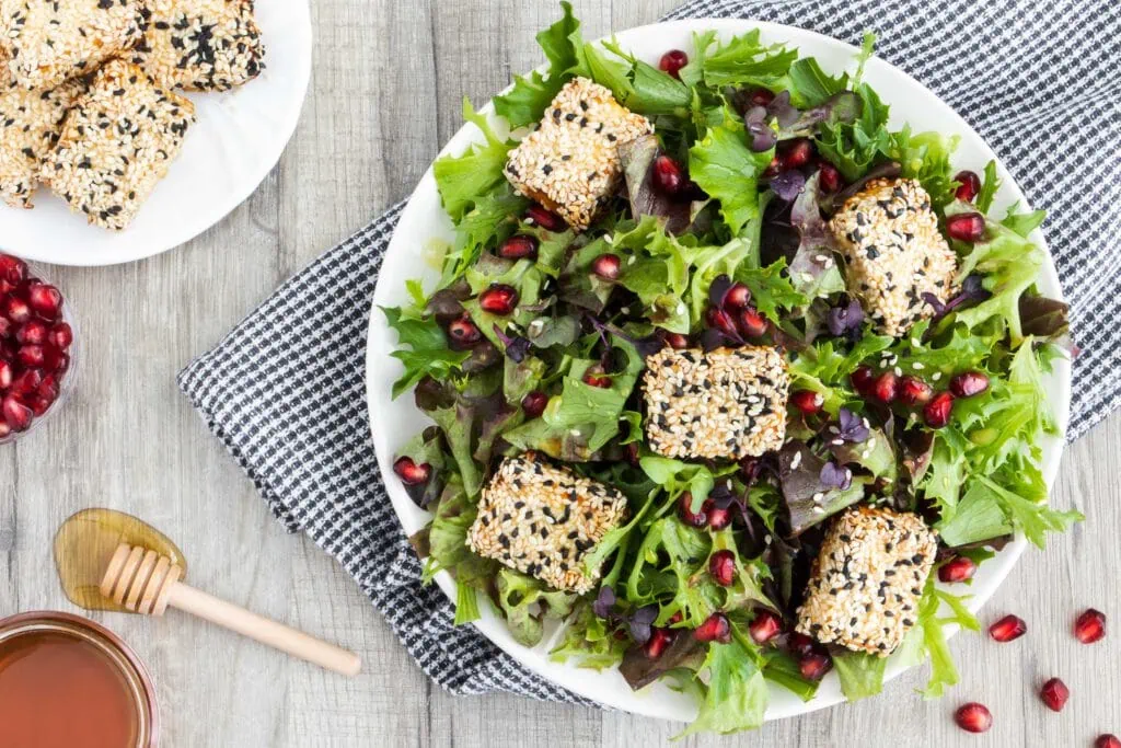 Salat mit Feta im Honig-Sesam Mantel angerichtet auf einem weißen Teller. Neben dem Salat liegen ein Honiglöffel, eine Schale mit Granatapfelkernen und ein kleiner Teller mit dem Feta im Honig-Sesam Mantel.