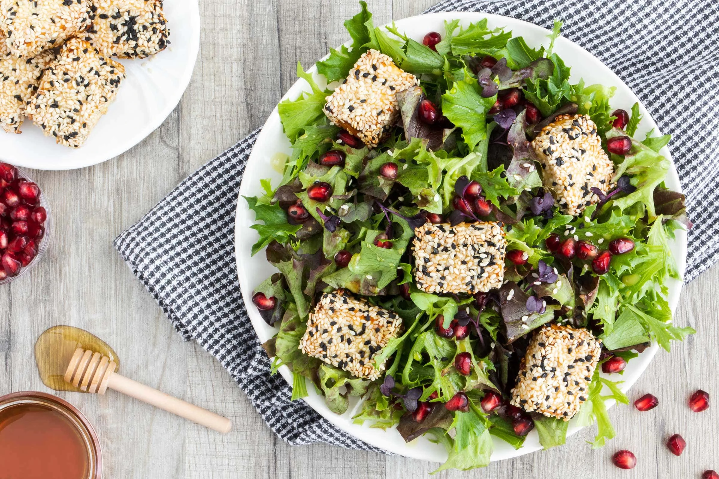 Salat mit Feta im Honig-Sesam Mantel angerichtet auf einem weißen Teller. Neben dem Salat liegen ein Honiglöffel, eine Schale mit Granatapfelkernen und ein kleiner Teller mit dem Feta im Honig-Sesam Mantel.
