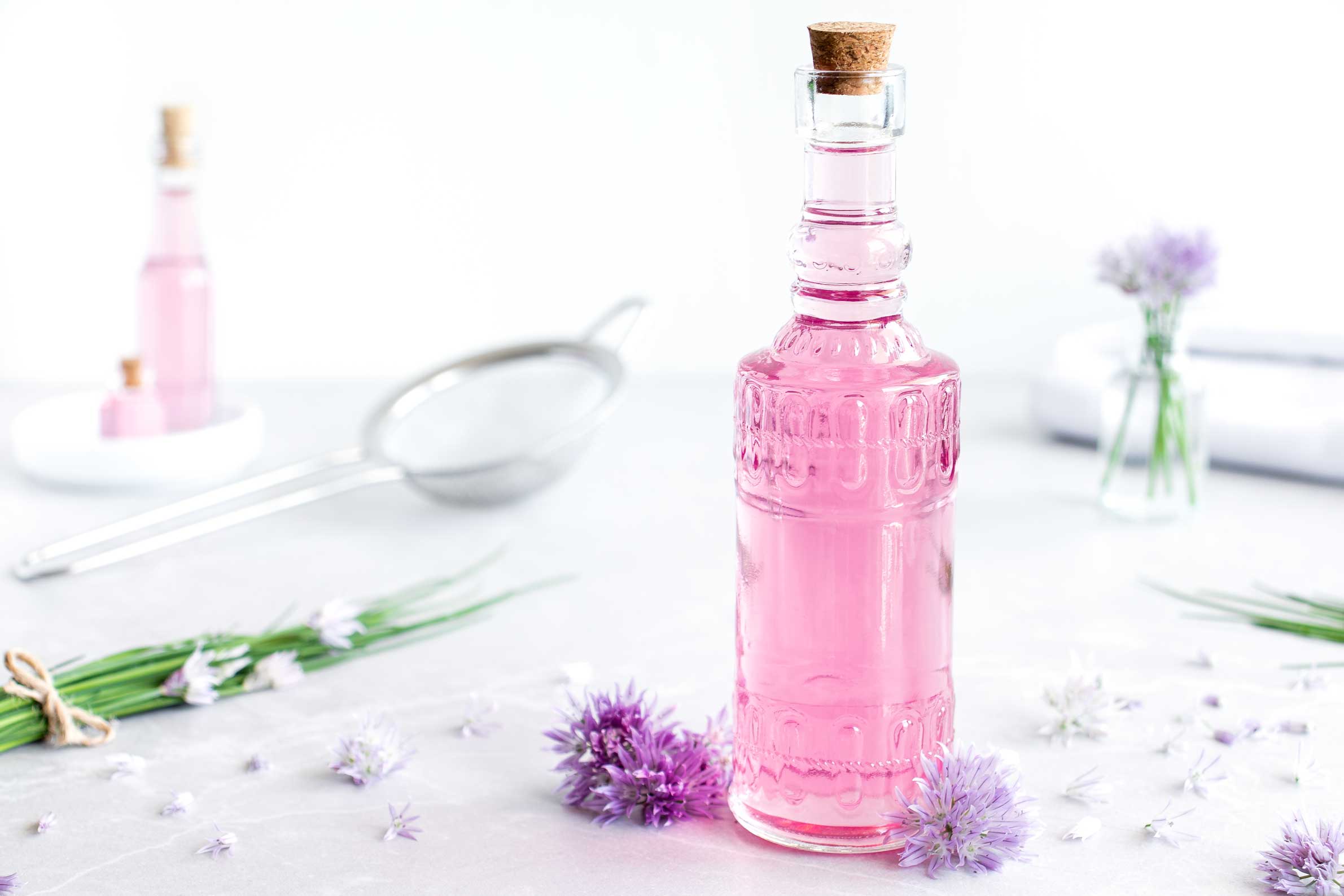 Schnittlauchblütenessig in einer Flasche auf einem weißen Untergrund. Neben dem Schnittlauchblütenessig liegen ein Bund Schnittlauch und mehrere Schnittlauchblüten.
