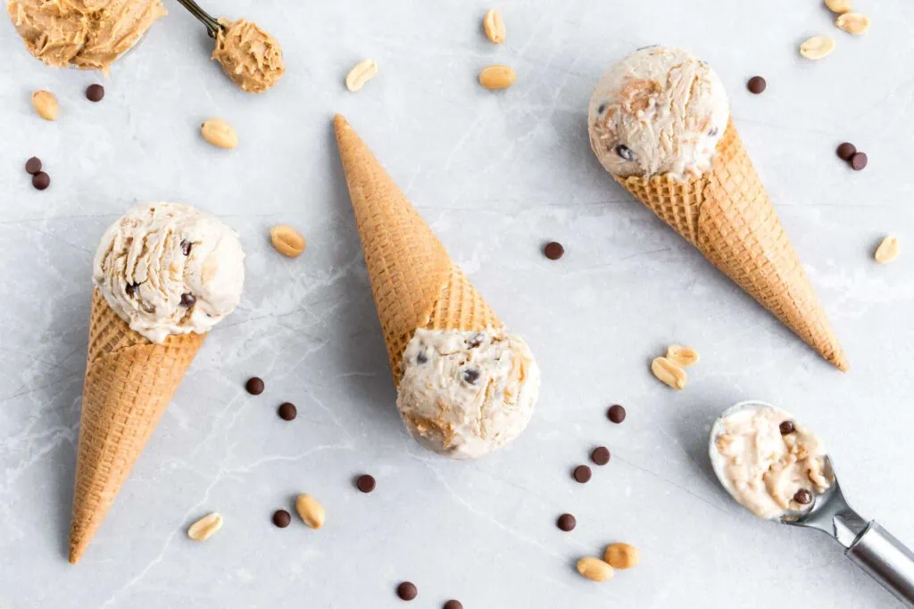 Erdnussbutter-Eis in Eiswaffeln. Neben dem Eis liegen mehrere Erdnüsse und Schokotröpfchen und ein Schälchen und ein Löffel mit Erdnussbutter.