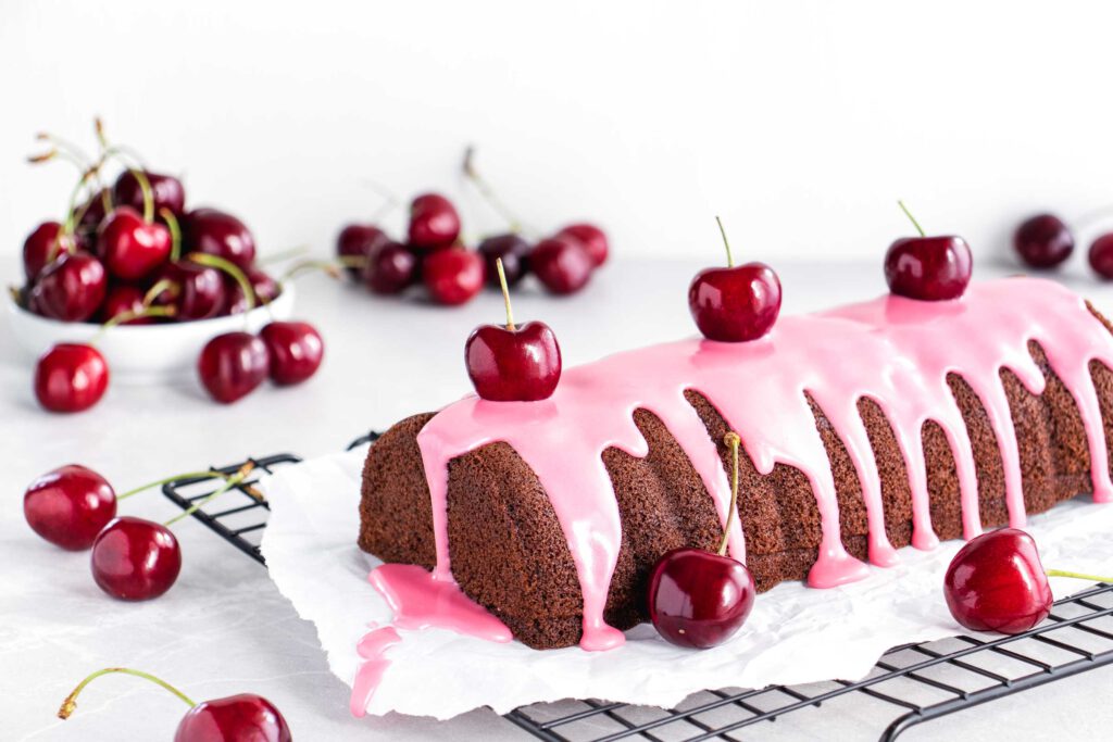 Schoko-Kirsch-Kuchen mit einer rosa Glasur aus Zuckerguss auf einem schwarzen Kuchengitter, verziert mit frischen Kirschen.