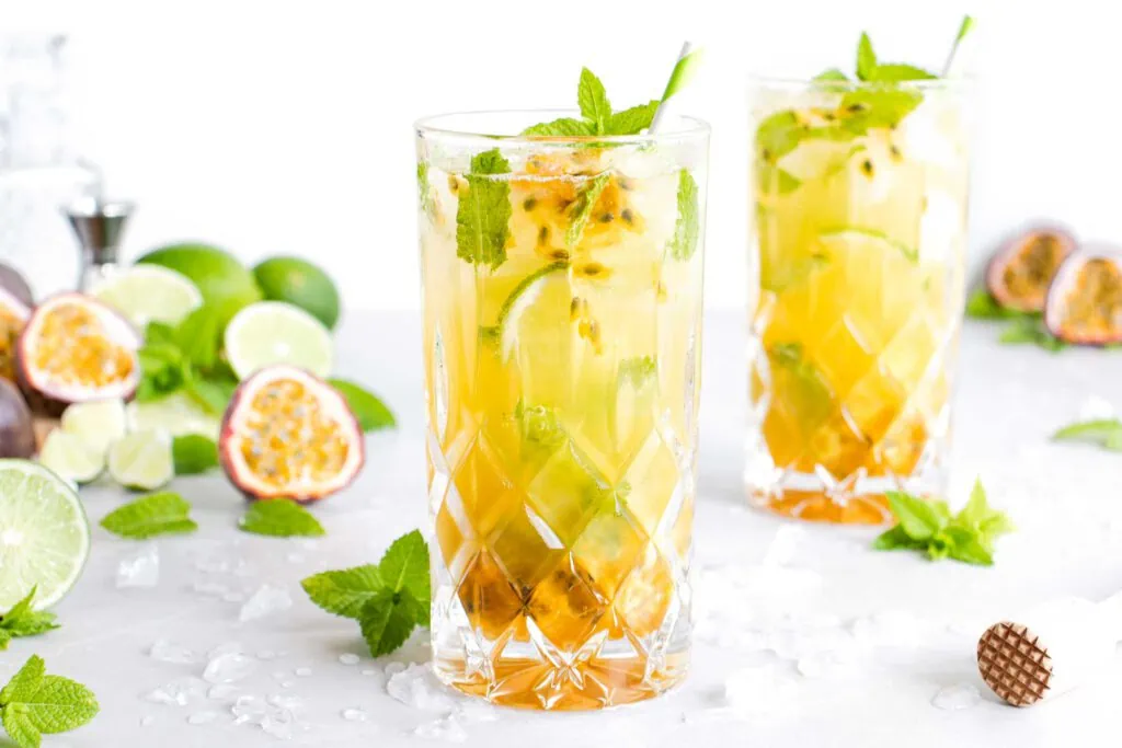 Maracuja Mojito in einem hohen Glas mit Crushed Ice, frischer Minze und Limettenstücken. Neben dem Cocktail liegen angeschnittene Passionsfrüchte und Limetten.