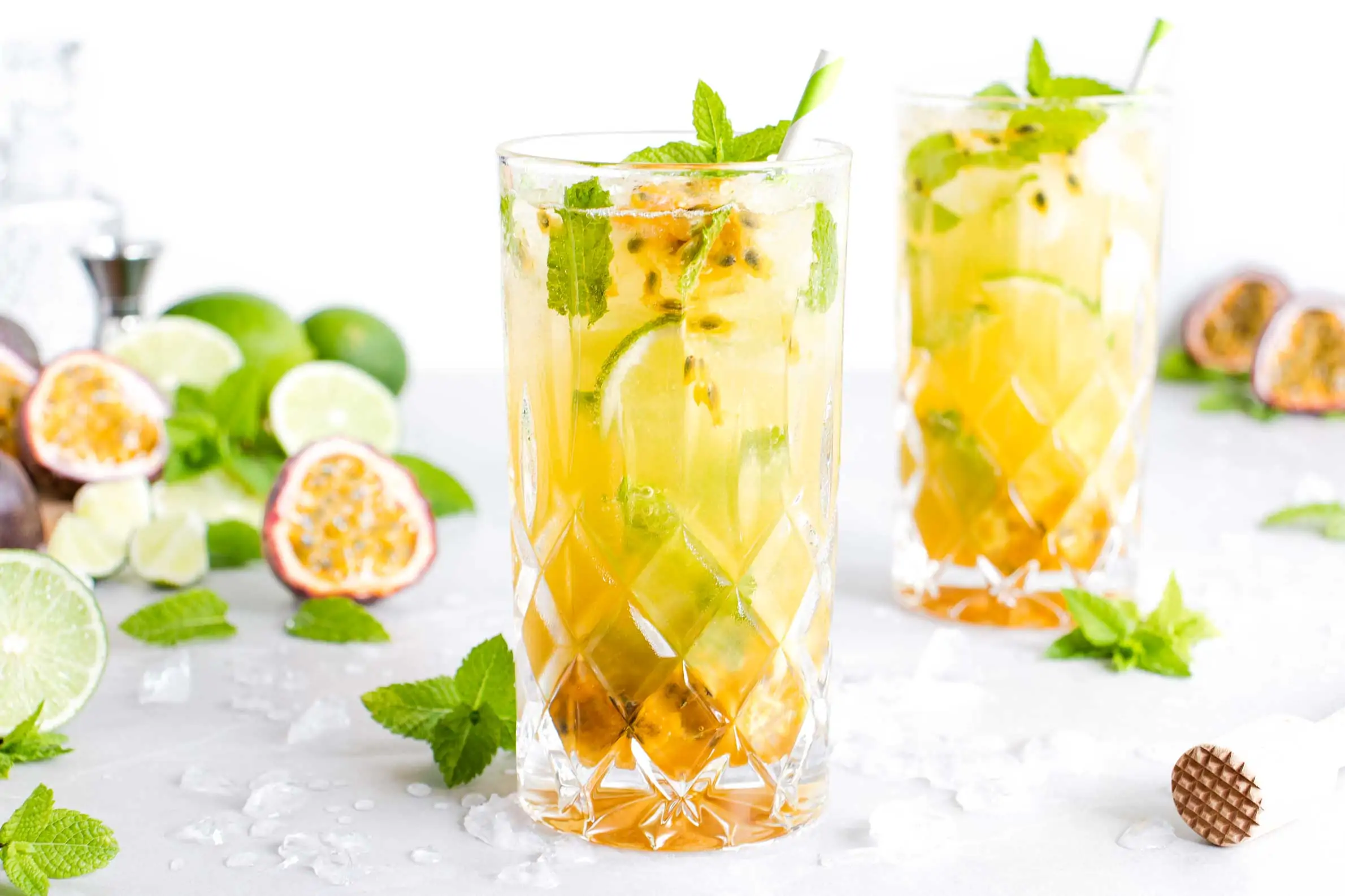 Maracuja Mojito in einem hohen Glas mit Crushed Ice, frischer Minze und Limettenstücken. Neben dem Cocktail liegen angeschnittene Passionsfrüchte und Limetten.