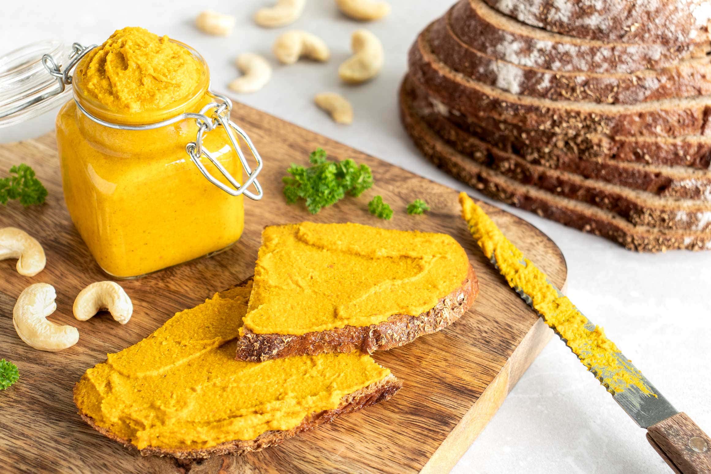 Kürbisaufstrich mit Cashews und Curry auf zwei Scheiben Brot. Im Hintergrund steht ein Glas mit dem gelben Brotaufstrich.