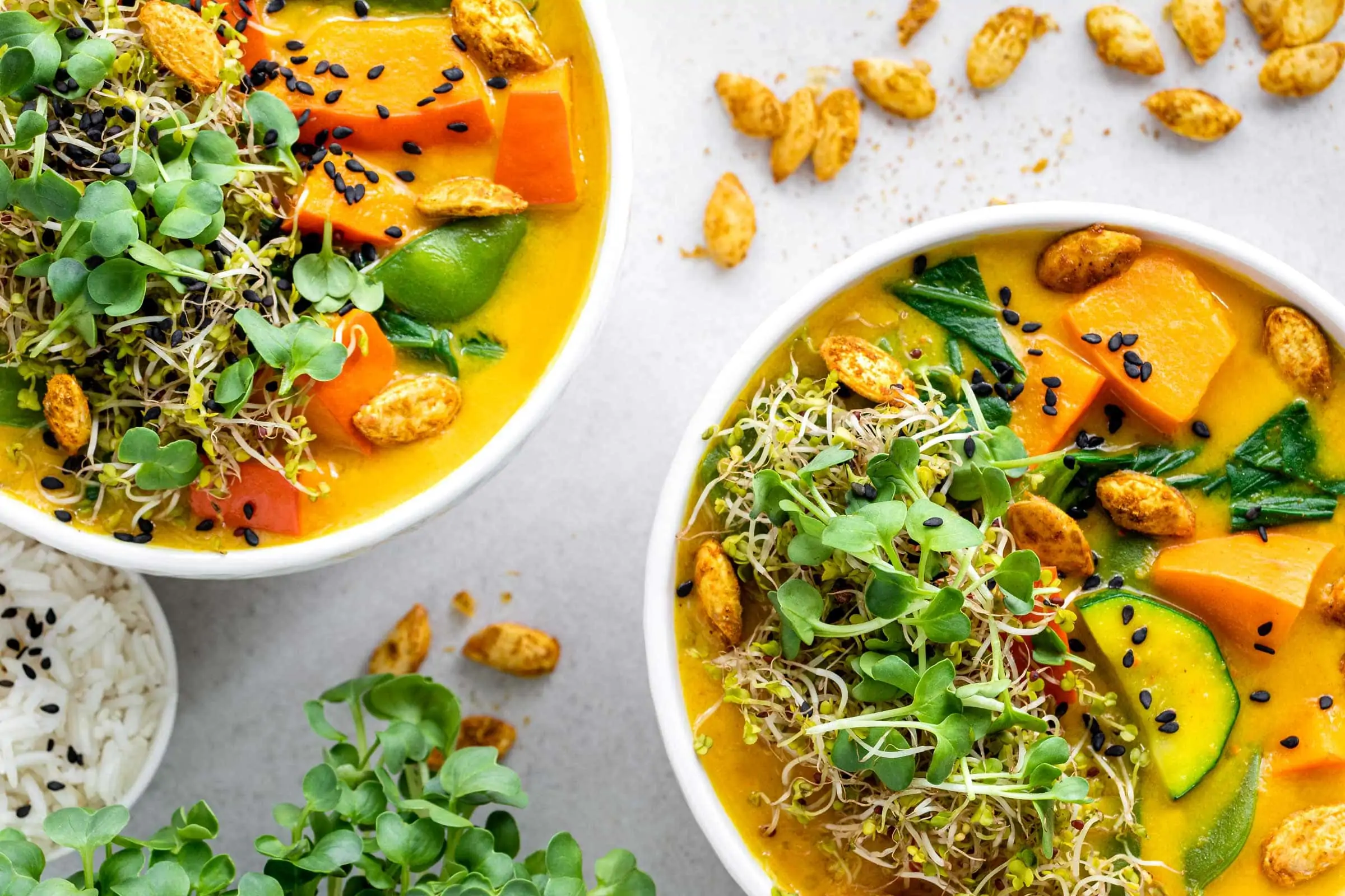 Kürbiscurry mit Kokosmilch in zwei weißen Schüsseln. Daneben steht eine kleinere Schüssel mit Reis.
